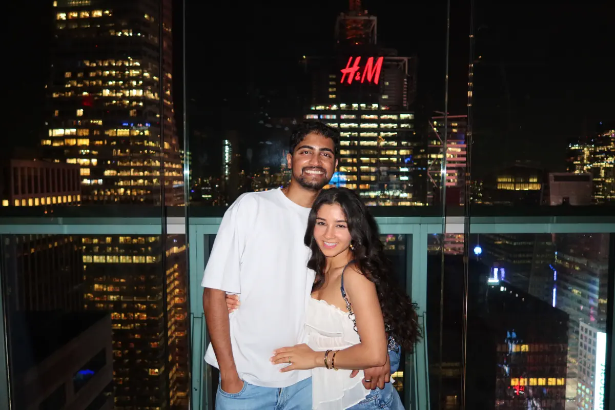 Naga and Lata on a rooftop with the city skyline lit up behind them