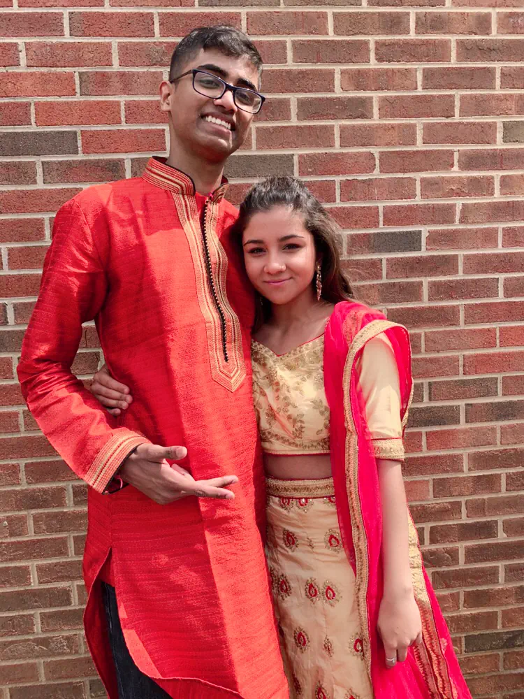 Naga and Lata dressed up in a red kurta and gold lehenga against a brick wall